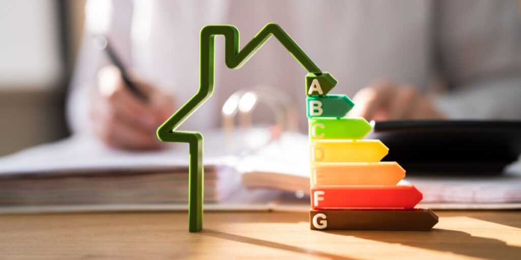 Green house-shaped energy efficiency model on a desk with a person writing in the background.
