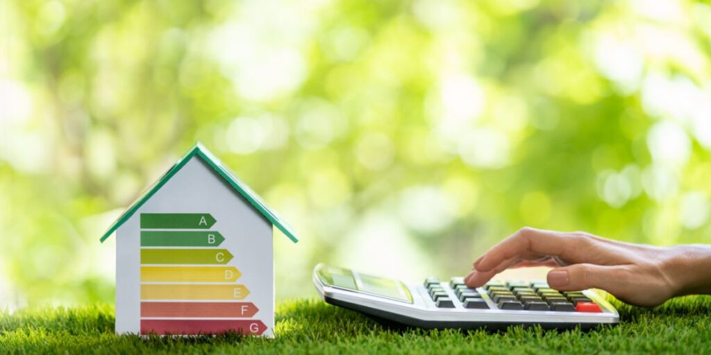 Hand pressing calculator beside a house-shaped energy efficiency model with a green-to-red scale.